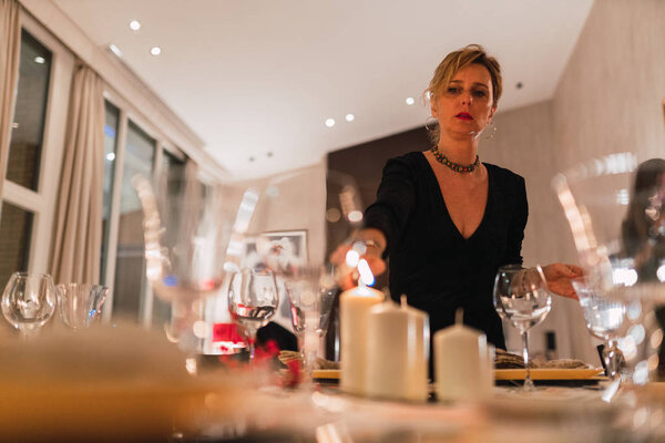 Снизу у взрослой элегантной женщины в черном платье зажигают свечи на столе, накрытом на праздничный ужин вечером
