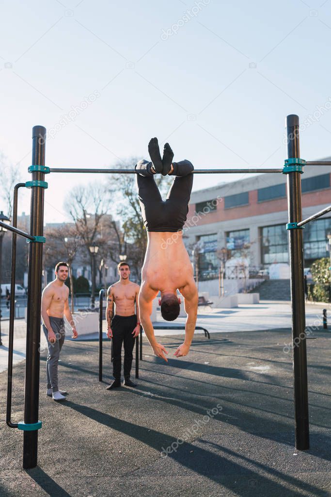 Back view of young sportive shirtless man hanging upside down on chin up bar in outdoor gym in sunny day