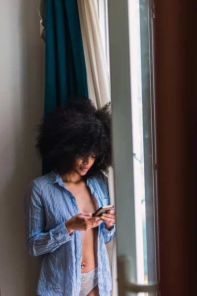 Joven encantadora mujer negra usando teléfono inteligente cerca de la ventana — Foto de Stock