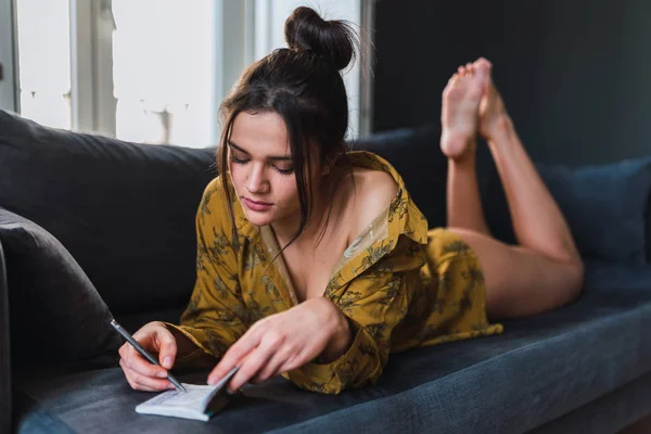 Młoda kobieta z długopis i Notes, leżąc na kanapie w pokoju — Zdjęcie stockowe