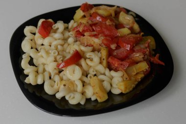 Beyaz arka planda izole edilmiş siyah bir tabakta haşlanmış sebzeli makarna.