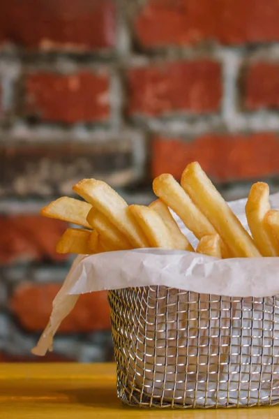 French fries in metal basket on yellow wooden table. Red brick backdrop — Stock Photo, Image