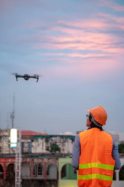 Young Asian engineer flying drone over construction site during sunset. Using unmanned aerial vehicle (UAV) for land and building site survey in civil engineering project.