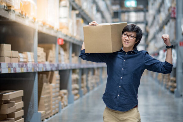 Young Asian happy man carrying cardboard box between row of shelves in warehouse, shopping warehousing or working pick and packing concepts