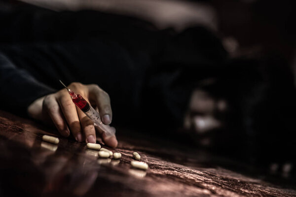 Junkie man lying on the floor while holding drug injection syringe and pills. Death drom drug overdose and addiction concept