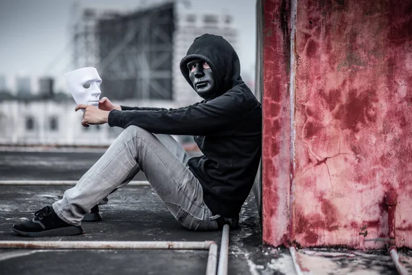 Mystery man wearing black mask and hoody jacket holding white mask sitting on rooftop of abandoned building, depression self destruction suicidal addiction massive depressive disorder concept