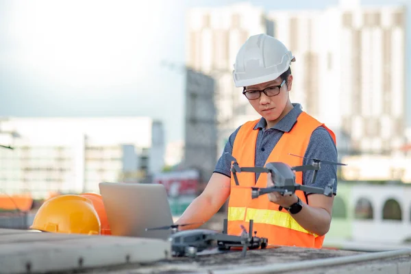 Young Asian man working with drone laptop and smartphone at construction site. Using unmanned aerial vehicle (UAV) for land and building site survey in civil engineering project.