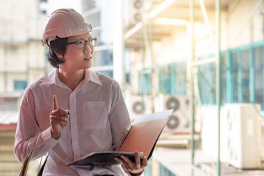 Genç Asyalı mühendis veya mimar inşaat alanında kişisel koruyucu ekipman kask giyen süre dosyaları tutan. Mühendislik, Mimarlık ve inşaat iş kavramları