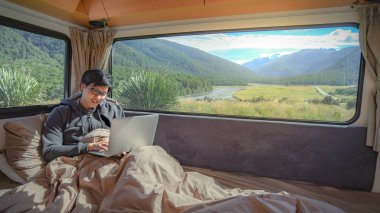 Genç Asyalı adam camper van pencereden doğal manzaralı, yol çelme takmak konsepti üzerinde dijital nomad ile yatakta dizüstü bilgisayar ile çalışma