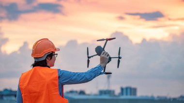 Genç Asyalı mühendis dron günbatımı sırasında inşaat alanında tutarak. İnsansız hava aracı (İHA) arazi için kullanarak ve inşaat mühendisliği proje sitesi anket oluşturma.