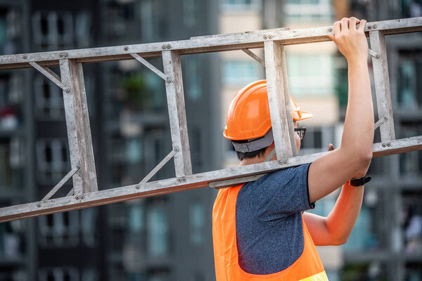 Young Asian maintenance worker with orange safety helmet and vest carrying aluminium step ladder at construction site. Civil engineering, Architecture builder and building service concepts