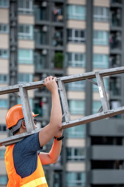 Young Asian maintenance worker with orange safety helmet and vest carrying aluminium step ladder at construction site. Civil engineering, Architecture builder and building service concepts