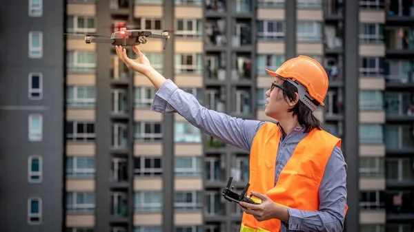 Young Asian engineer flying drone over construction site. Using unmanned aerial vehicle (UAV) for land and building site survey in civil engineering project.