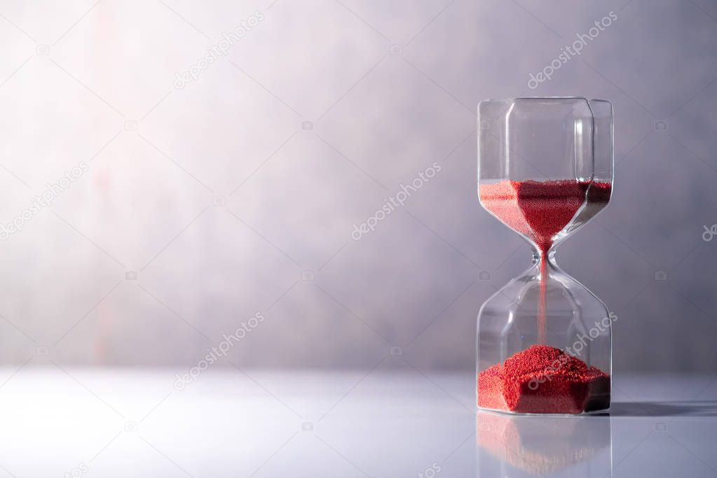 Red sand running through the shape of modern hourglass on white table.Time passing and running out of time. Urgency countdown timer for business deadline concept with copy space