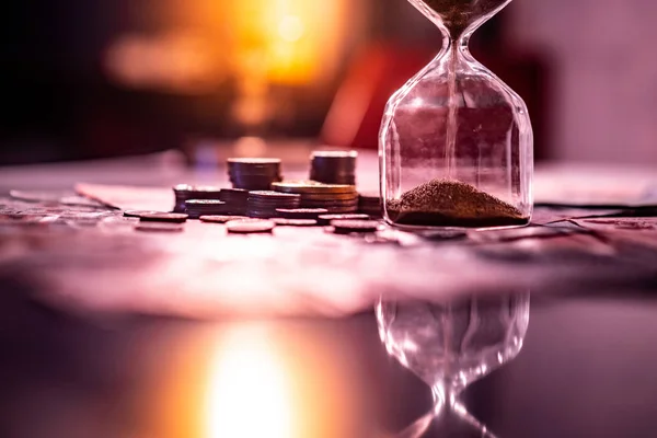 Sand running through the shape of hourglass on table with banknotes and coins of international currency. Time investment and retirement saving. Urgency countdown timer for business deadline concept