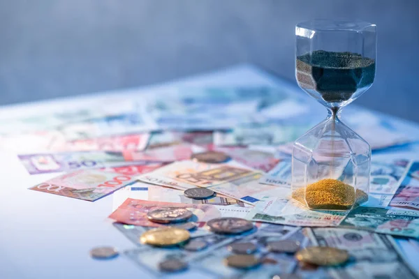 Sand running through the shape of hourglass on table with banknotes and coins of international currency. Time investment and retirement saving. Urgency countdown timer for business deadline concept