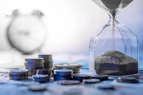 Sand running through the shape of hourglass on table with banknotes and coins of international currency. Time investment and retirement saving. Urgency countdown timer for business deadline concept