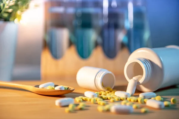 Pills spilling out of pill bottles with pharmaceutical spoon on wooden board. Table clock and test tube in the background. Prescription medicine and medical treatment concepts