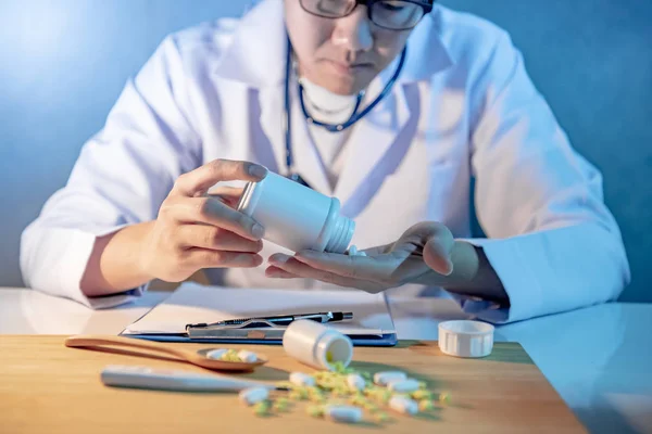 Male Asian doctor pouring pills from pill bottle to his hand. Doctor working with medicine paperwork clipboard on the desk in hospital clinic. Medical treatment and health care diagnostic concepts.