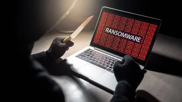 Male hacker threading laptop computer with knife showing on screen with red digital binary ransomware warning. Cyber attack and internet data security concept