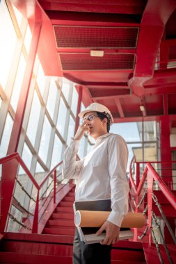 Küçük erkek mühendis veya mimar holding dosyaları ve giyen kask inşaat sahasında kırmızı merdiven üzerinde planları Asya. İnşaat Mühendisliği, Mimarlık ve inşaat kavramı bina