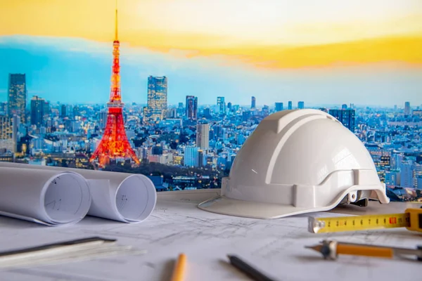 Architectural drawing plan with blueprint rolls, safety helmet and drawing tools on working table. Blurred Tokyo city in the background. Architecture and building construction concept