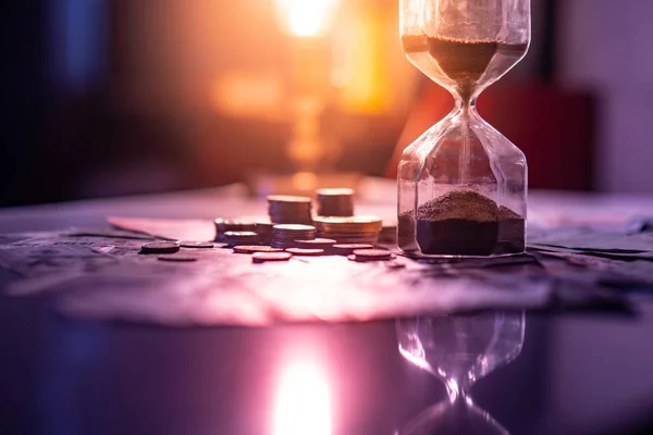 Sand running through the shape of hourglass on table with banknotes and coins of international currency. Time investment and retirement saving. Urgency countdown timer for business deadline concept