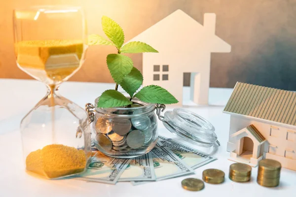 Real estate investment or property ladder. Home mortgage loan rate. Saving money for future concept. Plant growing out of coins in glass jar with dollar banknotes, hourglass and house model on table