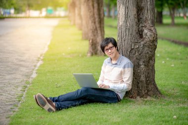 Küçük Asya mutlu adam bahçede çimlerin üzerine beyaz bilgisayar dizüstü oturan kullanarak. Erkek üniversite öğrenci açık rahatlatıcı. Eğitim ve teknoloji kavramları