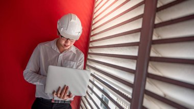 Genç Asyalı erkek mühendis veya mimar giyiyor koruyucu kask ve kırmızı ile dizüstü bilgisayarla çalışma gözlük arka planda duvar. Mimarlık, mühendislik ve inşaat kavramlar bina