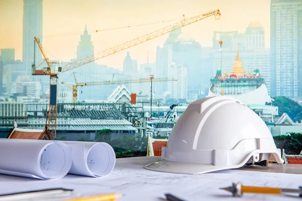 Mimari çizim planı planı ile rulo, çalışma tablosu üzerinde güvenlik kask ve çizim araçları. Bulanık vinç ve inşaat site Bangkok içinde belgili tanımlık geçmiş. Bina inşaat kavramı