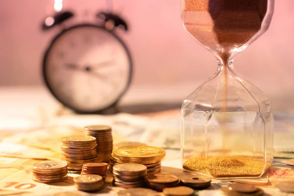 Sand running through the shape of hourglass on table with banknotes and coins of international currency. Time investment and retirement saving. Urgency countdown timer for business deadline concept