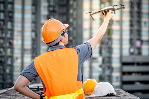 Young Asian engineer man working with drone laptop and smartphone at construction site. Using unmanned aerial vehicle (UAV) for land and building site survey in civil engineering project.