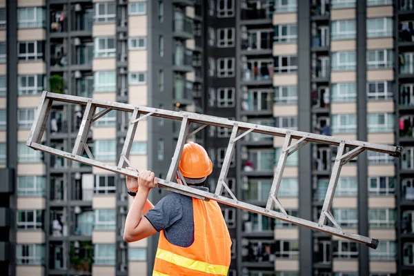 Young Asian maintenance worker man with orange safety helmet and vest carrying aluminium step ladder at construction site. Civil engineering, Architecture builder and building service concepts