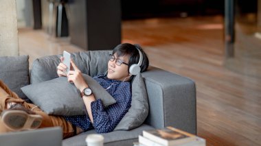 Mobil uygulama müzik dinlerken, yalan ve serbest zaman sırasında kanepede rahatlatıcı için Smartphone kullanan Genç Asyalı adam. Yaşam alanı kavramı kentsel yaşam tarzı