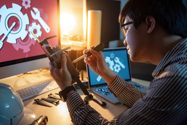 Asian technical engineer using screwdriver for repairing drone with computer and tools on desk. Male technician fixing or maintenance drone. Unmanned aerial vehicle (UAV) photography concept