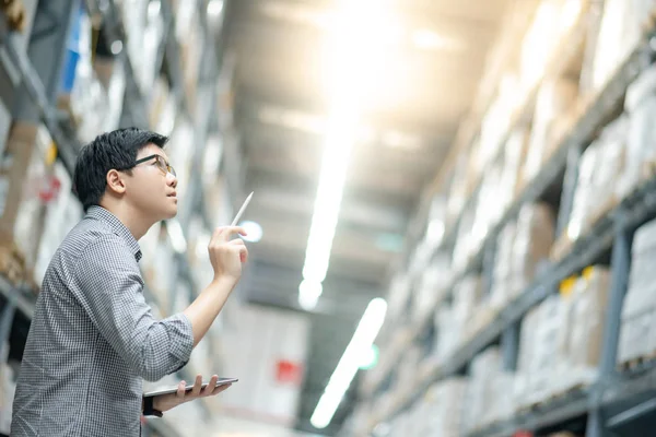 Genç Asyalı işçi, dijital tablet ve kalem kullanarak depodaki raflardaki karton kutularda ürün stokluyor. Fiziksel envanter sayımı konsepti