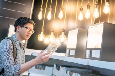Young Asian man choosing package of electrical light bulb in the store. Furniture home decoration shopping concept clipart