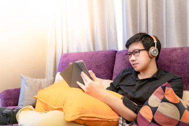 Oturma odasında kanepede dijital tablet ve kalem kullanarak gözlük ve kulaklık ile genç Asya mutlu adam. Ev yaşam yaşam tarzı modern elektronik aygıtı kavramı ile