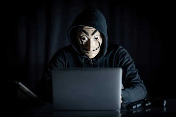 男性连帽衫黑客戴着神秘面具使用笔记本电脑坐在桌子上的数字平板电脑和枪 匿名社交屏蔽 勒索网络攻击或互联网安全概念 — 图库照片