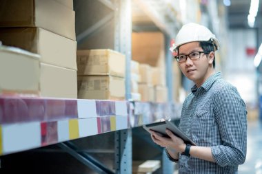 Güvenlik kaskı ve gözlük takan genç Asyalı işçi dijital tablet ve kalem kullanarak depodaki raflardaki karton kutularda ürün stokluyor. Fiziksel envanter sayımı konsepti