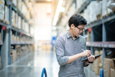 Genç Asyalı işçi, dijital tablet ve kalem kullanarak depodaki raflardaki karton kutularda ürün stokluyor. Fiziksel envanter sayımı konsepti