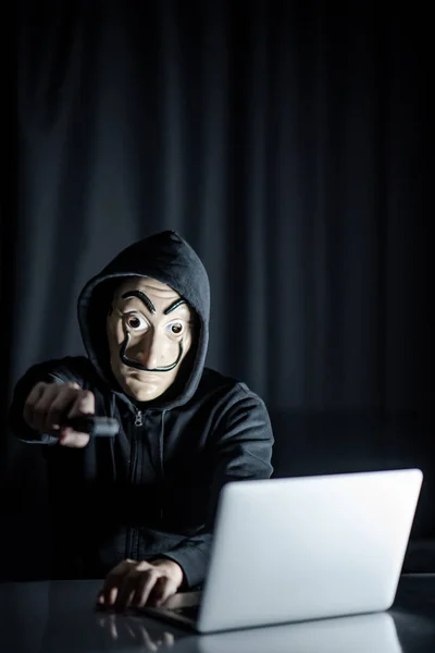 男性连帽衫黑客戴着神秘面具手持枪 而坐在桌上的笔记本电脑 勒索网络攻击或互联网安全概念 匿名小偷犯下的罪行 — 图库照片