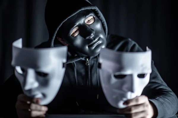 Mystery hoody man wearing black mask holding two white masks in his hand. Anonymous social masking. Major depressive disorder or bipolar disorder. Halloween concept