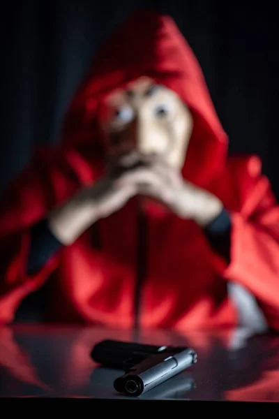 Mystery man in red hoodie sitting with black gun on the table. Anonymous thief or robber committing a crime. Weapon and violence concepts