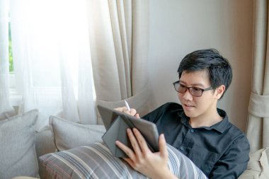 Koltuk veya kanepe oturma odasında otururken dijital tablet ve kalem kullanarak gözlük ile genç Asya iş adamı. Ev yaşam yaşam tarzı modern elektronik aygıtı kavramı ile