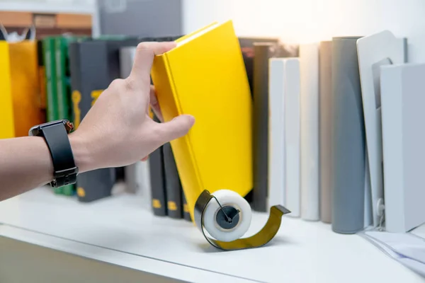 Male hand choosing and picking yellow book from white bookshelf in public library. Education research and self learning in university life concepts