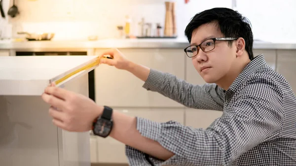 Young Asian man using tape measure for measuring granite countertops on modern kitchen counter in showroom. Shopping furniture for home improvement. Interior design concept