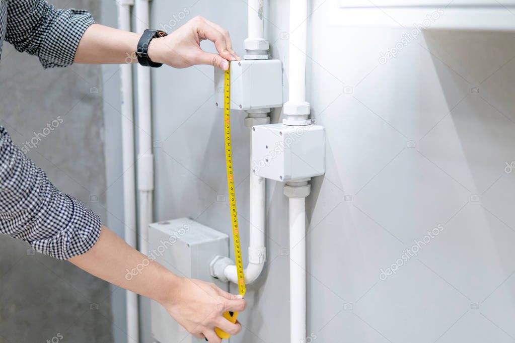 Male hand electrician using tape measure for measuring dimension of electrical conduit and electrical box on the wall. Electrical fitting installation and building construction concepts