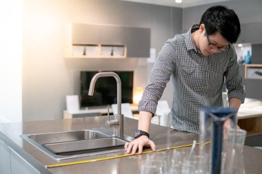 Genç Asyalı adam sergi salonundaki mutfak tezgahında granit tezgahları ölçmek için mezura kullanıyor. Ev dekorasyonu için mobilya alışverişi. İç tasarım kavramı